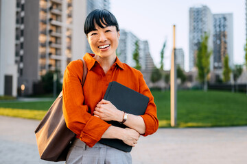 Asian woman in orange shirt smiling while holding a laptop bag in an urban park on a sunny day