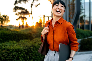 Joyful Asian woman enjoying an evening stroll in the city park during sunset, holding a laptop and...