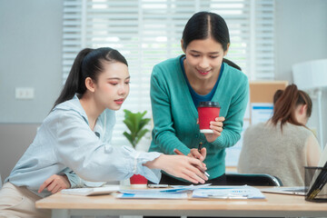 Three Asian women collaborate at shared office desk filled with documents, charts, coffee. One...