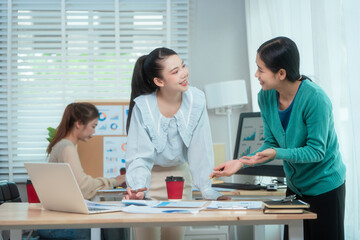 Fototapeta premium Three Asian women collaborate at shared office desk filled with documents, charts, coffee. One leads the discussion, another points at data, third listens attentively, showing productive teamwork.