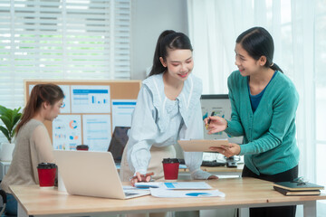 Three Asian women collaborate at shared office desk filled with documents, charts, coffee. One...