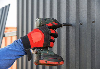 A gloved construction worker uses a drill driver to drive a screw into a dark corrugated metal...