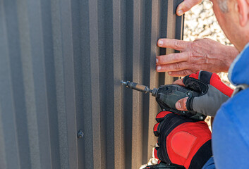 A gloved construction worker uses a drill driver to drive a screw into a dark corrugated metal...