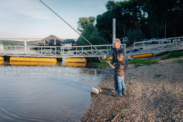 Grandfather teaching grandson how to fish on river bank
