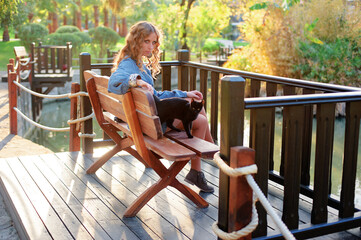 A young woman enjoys a tranquil moment with her black cat on a riverfront bench amidst lush gardens.