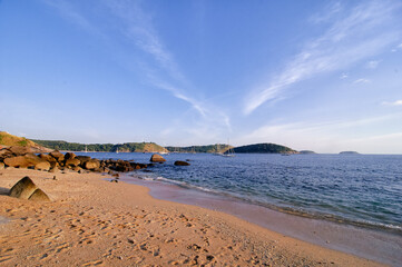 Tranquil coastal view featuring sandy beach, scattered rocks, and sailboats on calm waters. The serene scene is complemented by nearby green hills and a vast blue sky.