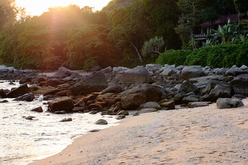 Serene beach scene featuring a sunset over a rocky shoreline bordered by lush tropical greenery....