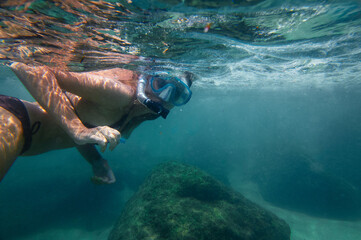 A woman wearing snorkeling gear explores a vibrant underwater world, swimming above rocks immersed...