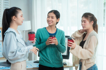 Three Asian women collaborate at shared office desk filled with documents, charts, coffee. One...
