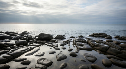 Obraz premium Dark rocks form natural pools at the edge of a calm ocean under a cloudy sky