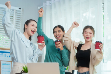 Three Asian women collaborate at shared office desk filled with documents, charts, coffee. One...