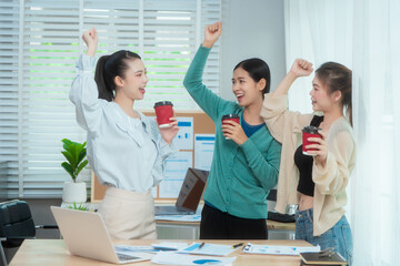 Three Asian women collaborate at shared office desk filled with documents, charts, coffee. One...
