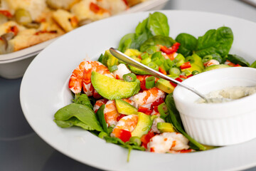 A vibrant salad featuring succulent shrimp, creamy avocado, and crisp lettuces, all served on a white plate. A side of creamy dressing in a bowl.