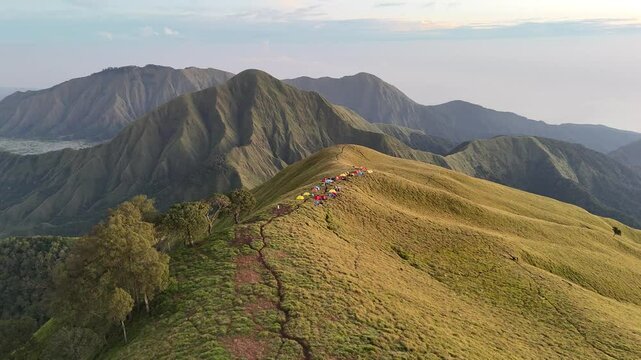 A stunning, static landscape view of the Mount Rinjani volcano from the crater rim campsite, with colorful hiker tents in the foreground under a clear blue sky on Lombok, Indonesia.