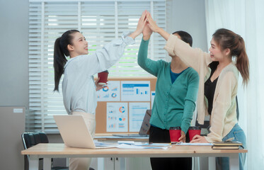 Three Asian women collaborate at shared office desk filled with documents, charts, coffee. One...
