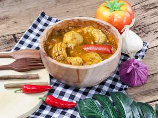 A bowl of spicy Indonesian seblak, featuring fish balls, corn, chili, and vegetables in a rich, savory broth. Surrounded by fresh garlic, onions, and chili peppers, the dish exudes vibrant flavors.