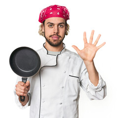 Young Caucasian male chef with frying pan smiling cheerful showing number five with fingers.