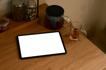 Digital tablet with blank screen on kitchen table beside glass teapot and cup of tea