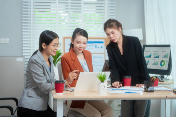 Three Asian women in formal suits collaborate at a desk, discussing documents in a modern office....
