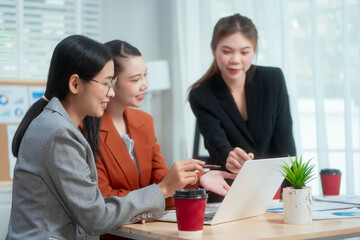 Three Asian women in formal suits collaborate at a desk, discussing documents in a modern office....