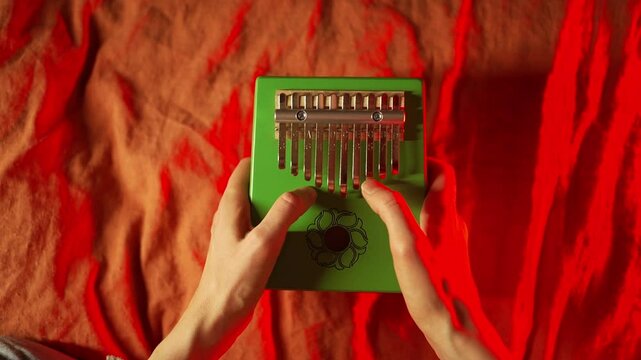 White-skinned musician's hands delicately playing green kalimba with floral decoration, resting on vibrant wrinkled red and orange fabric surface