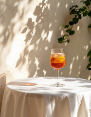 A vibrant Aperol Spritz, garnished with an orange slice, sits on a white tablecloth-covered outdoor table, illuminated by dappled sunlight and shadows from lush green foliage.