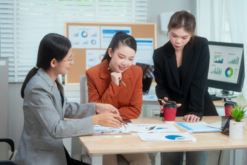 Three Asian women in formal suits collaborate at a desk, discussing documents in a modern office....