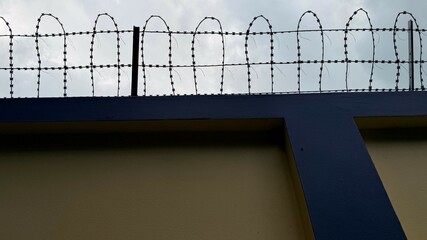Barbed wire strands sit atop a concrete wall under a cloudy, overcast sky