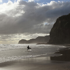 Emerging from the waves at Piha Beach, a lone surfer carries their board onto the dark sand, backed...