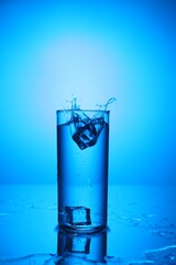 Fresh water with ice cubes splashing out of glass on mirror table against blue gradient background