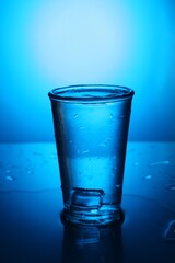 Fresh water with ice cube in glass on mirror table against blue gradient background. Backlit
