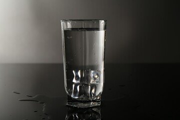 Fresh water with ice cubes in glass on mirror table against gray background