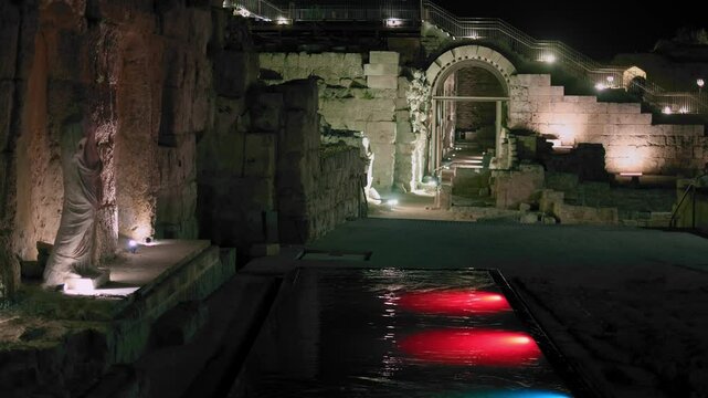 archaeological site in old caesarea israel at night. modern pool with water on the site of old antique bath with old statue near the pool and perspective on old arches and stairs. color illumination