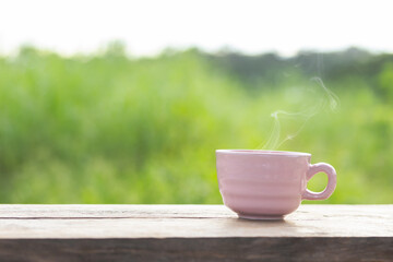 Hot coffee cup on wooden table in the garden, green leaves blurred as background, pink coffee cup