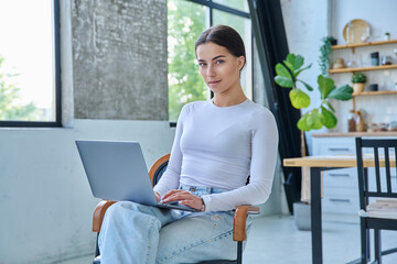 Portrait of young woman sitting on chair with laptop looking at camera, in home interior