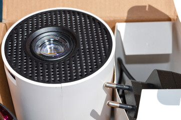 close-up of a hand holding a mini projector inside packaging box, visible power plug and lens...