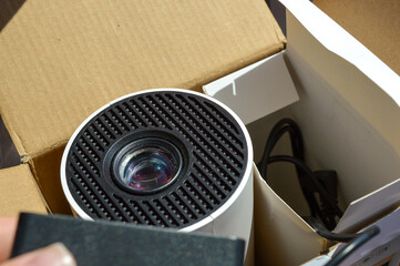close-up of a hand holding a mini projector inside packaging box, visible power plug and lens...