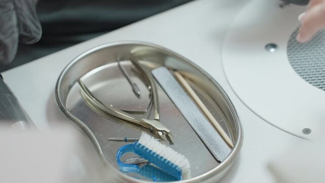 Close-up of nail technician wearing black glove selecting manicure tool from stainless tray containing various nail care instruments including buffer, stick, brush and clipper beside customer hand