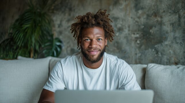 A joyful young man with dreadlocks is sitting on a couch, working on his laptop in a relaxed home environment, portraying a balance of work and enjoyment in daily life.