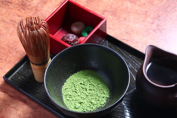 A traditional Japanese matcha tea set featuring a bowl of vibrant green tea powder, a bamboo whisk, and a small pitcher is artfully arranged on a dark tray.