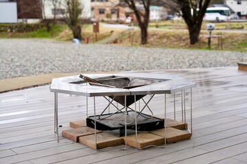 A modern outdoor fire pit or portable BBQ grill set up on a wooden deck with stone pavers underneath, ready for a camping trip or backyard gathering, in a gravel-covered outdoor space.