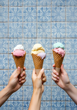 Three hands holding artisanal gelato cones with dual pastel flavors in front of a vintage blue-and-white tiled wall.