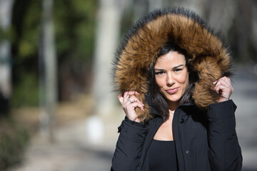 Portrait of a young, beautiful, dark-haired woman, looking at the camera, wrapping herself up with the fur collar of her coat, on a cold and sunny day. Concept winter, cold, autumn, weather.