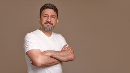 A confident man in his 50s, stands with arms crossed, wearing a white shirt against a plain brown...