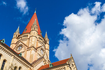 Obraz premium Stunning view of the historic St. Francis of Assisi Church in Vienna, Austria, showcasing its Romanesque Revival architecture under a bright blue sky with dramatic white clouds.