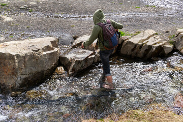 person with a backpack crosses a rocky stream in a natural landscape. Wearing a hoodie, they...