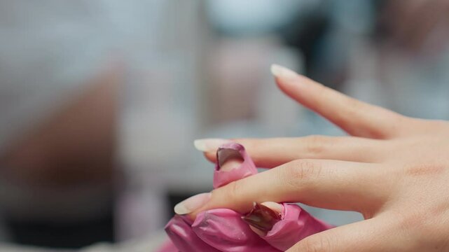 Close up view of nail technician wearing pink gloves carefully applying hardener with brush to client fingernail during manicure session, with soft blur background emphasizing detail and precision