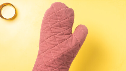 A pink quilted oven mitt shown against a bright yellow background, ideal for kitchen safety and heat protection, with a glass cup slightly blurred in the corner