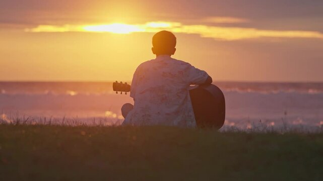A young man sits and plays guitar on a tropical beach at sunset. A happy man enjoys the sunset while playing guitar on the beach