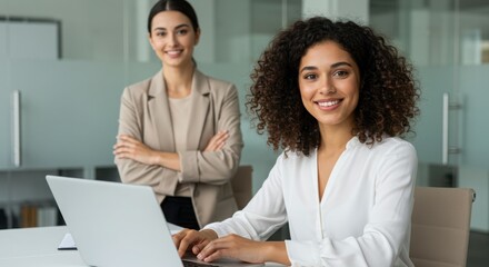 Obraz premium Two smiling businesswomen in office with laptop and arms crossed behind